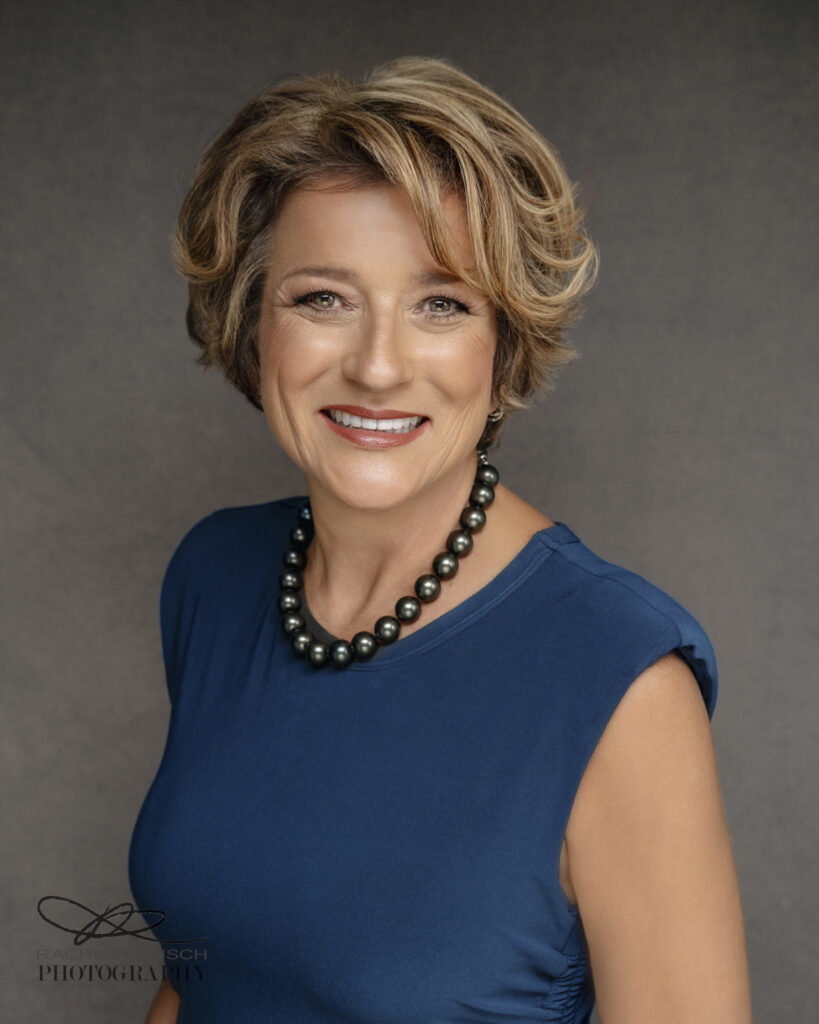 Professional headshot of a woman with short blonde hair in a navy top and pearl necklace